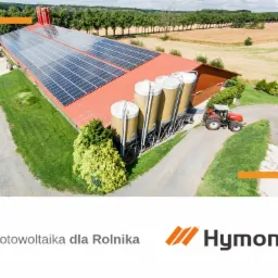 Widok z góry na duży budynek gospodarczy z panelami słonecznymi na dachu, silosami i czerwonym traktorem obok, otoczony zielenią i polami.