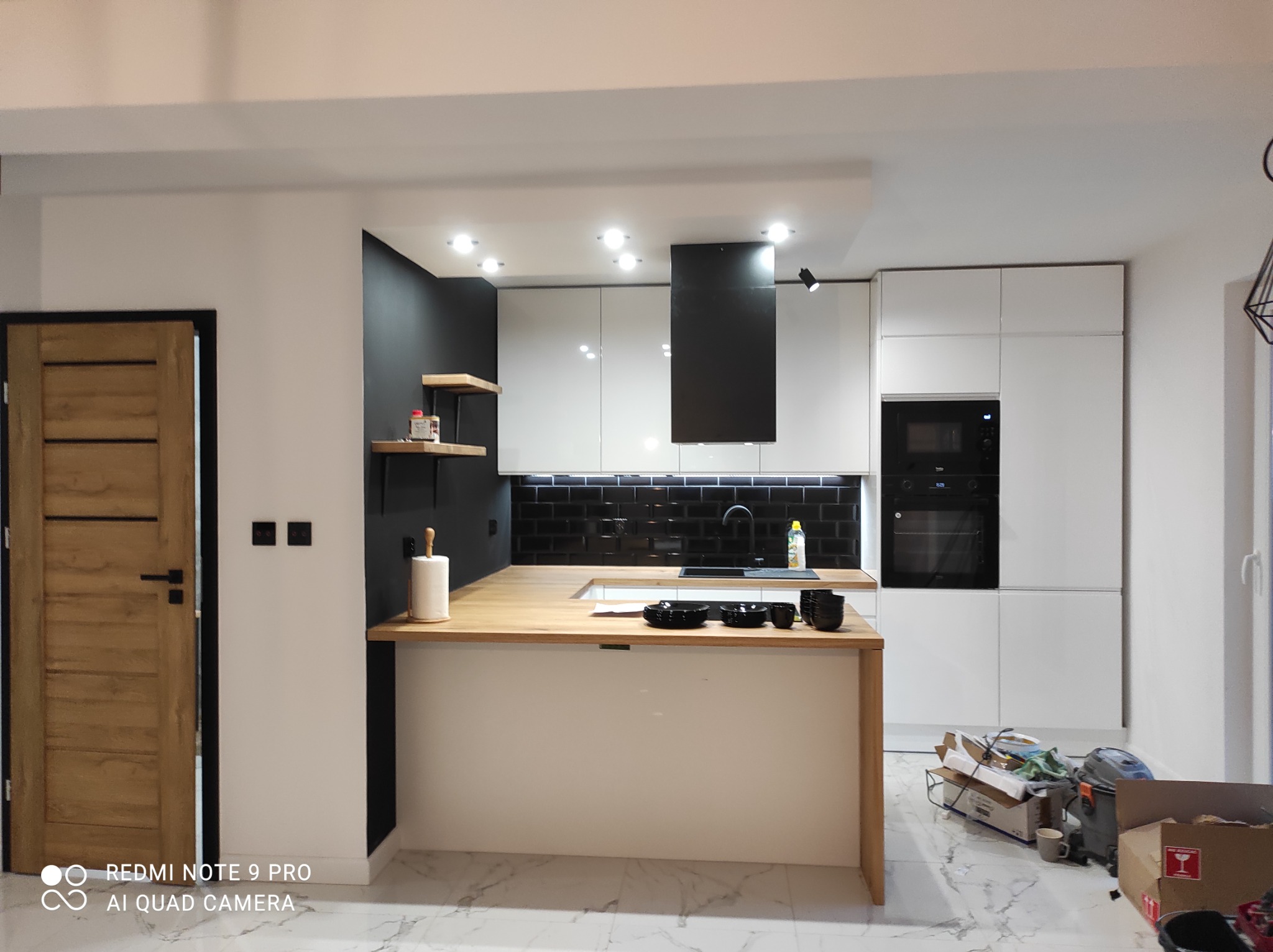 Nowoczesna kuchnia z białymi szafkami, czarnym panelem nad blatem i drewnianym blatem, widoczne czarne miseczki na wyspie kuchennej, oraz kartony po sprzętach w tle.