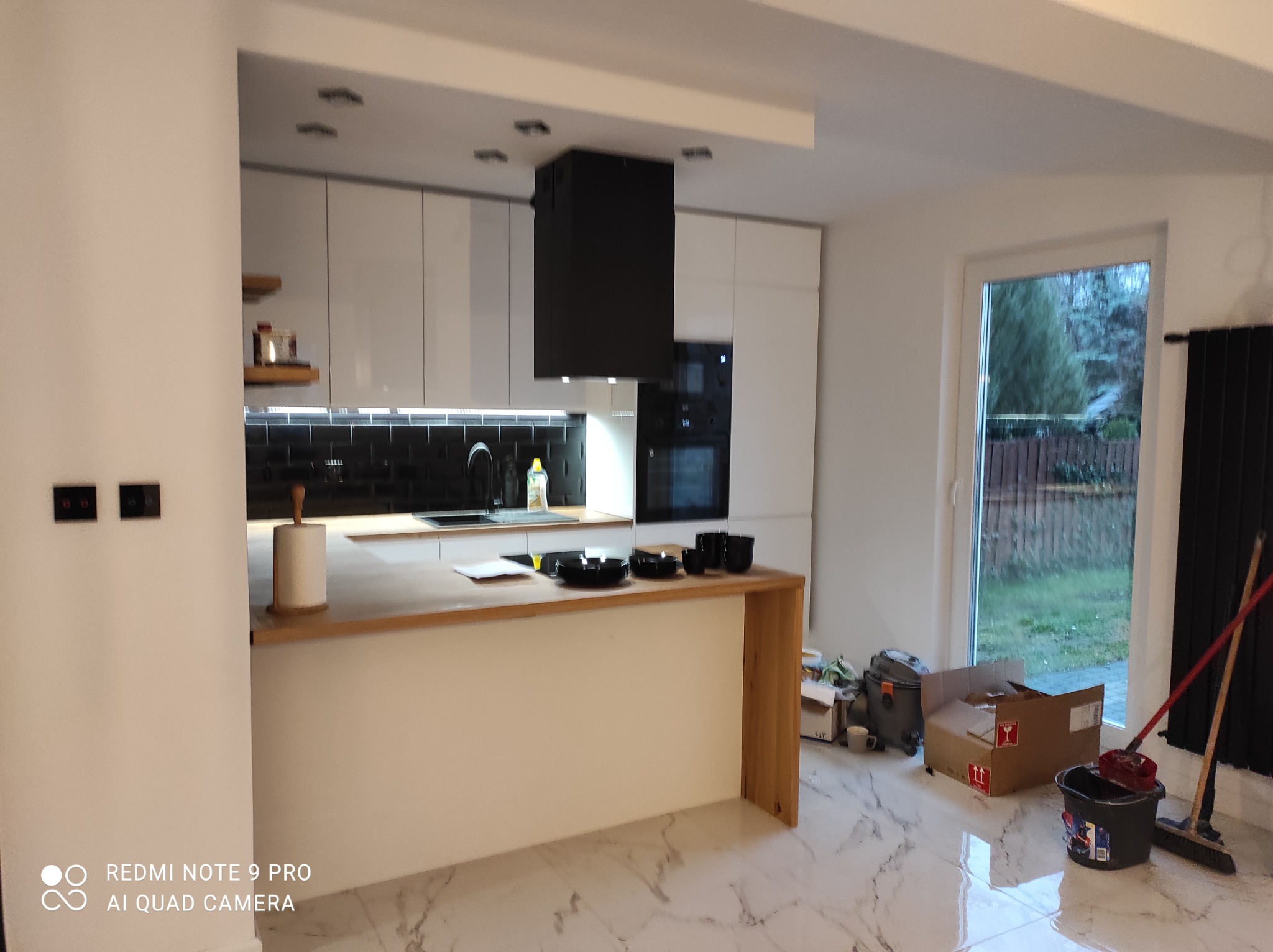 Nowoczesna kuchnia z białą zabudową, czarnym okapem i wyspą z drewnianym blatem, na marmurowej podłodze widoczne ślady po remoncie.