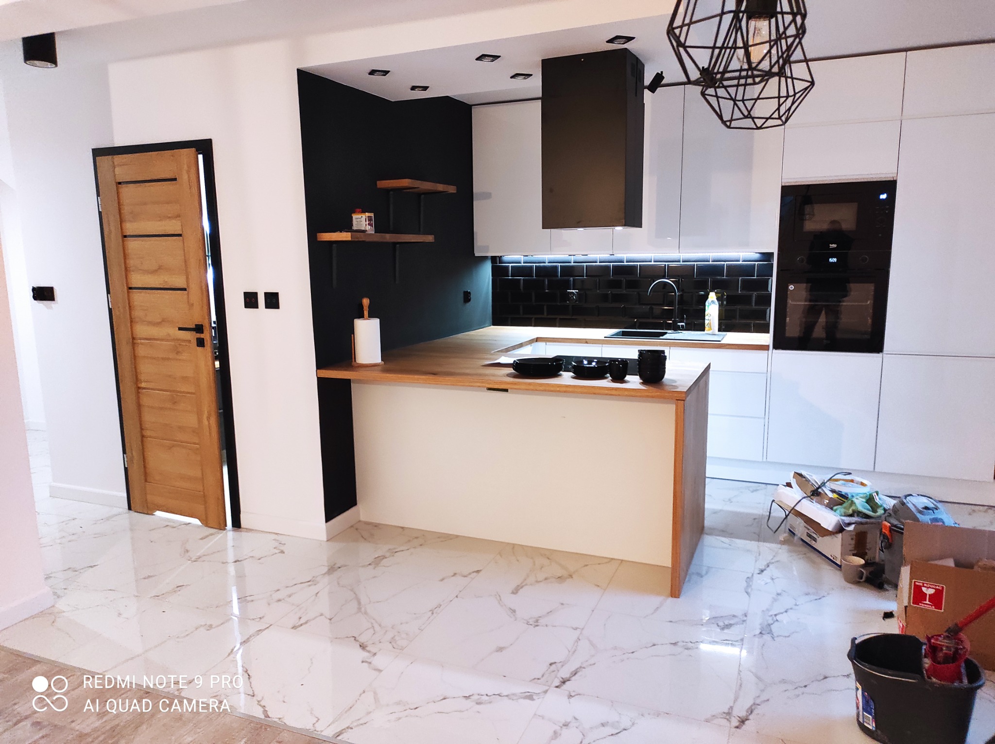Nowoczesna kuchnia z wyspą, czarne płytki nad blatem, białe szafki, drewniany blat i otwarte półki, widoczne kartony i wiadro po remoncie, marmurowe płytki podłogowe.