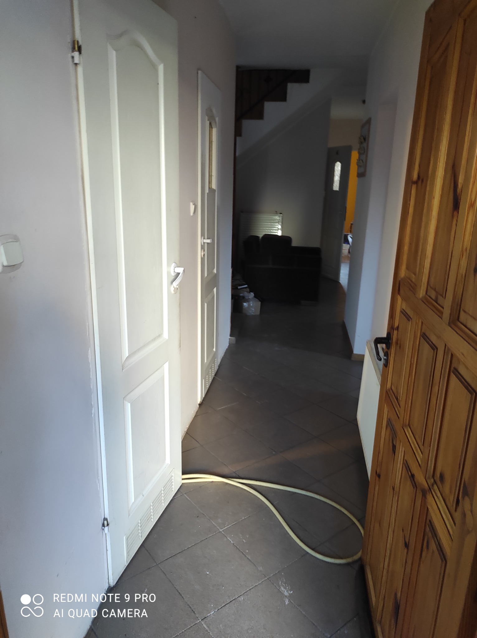 Otwarta, biała, wewnętrzna ościeżnica drzwiowa w Darłowie, z widocznym fragmentem podłogi z szarych płytek i żółtym wężem leżącym na podłodze. W tle widoczne schody i dalsza część mieszkania.