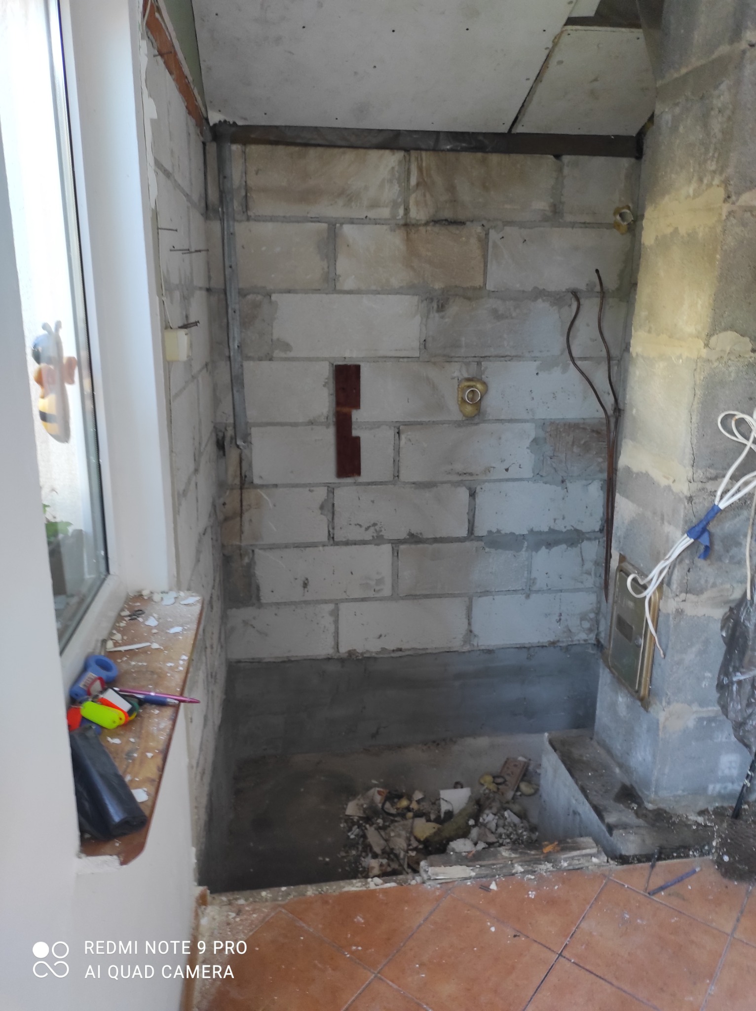 Wnętrze łazienki w trakcie demontażu, widoczne surowe ściany z bloczków, fragment okna z parapetem pokrytym drobnymi śmieciami, oraz fragment podłogi z brązowych płytek.