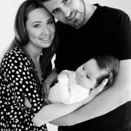 Czarno-białe zdjęcie młodej rodziny: uśmiechnięta kobieta w sukience w grochy, mężczyzna w czarnym T-shircie trzymający niemowlę na rękach, jasne tło.