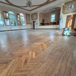 Sala balowa z parkietem ułożonym w jodełkę przed cyklinowaniem, widoczny odkurzacz przemysłowy i ozdobne piece kaflowe.