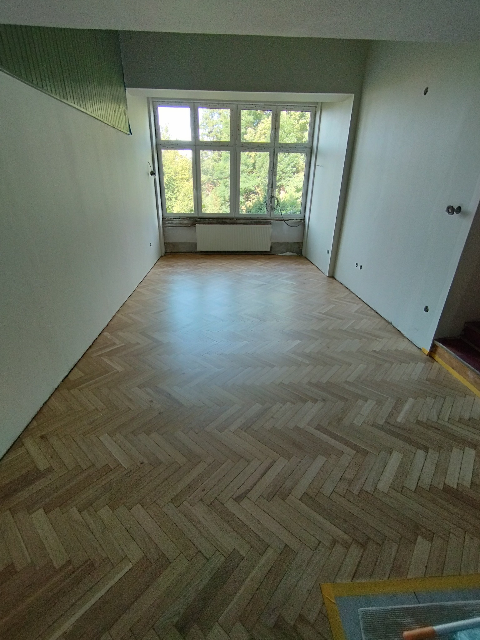 Jasny parkiet ułożony w jodełkę w pustym pokoju z dużym oknem i grzejnikiem, ściany w trakcie malowania, widoczne ślady po gniazdkach elektrycznych.