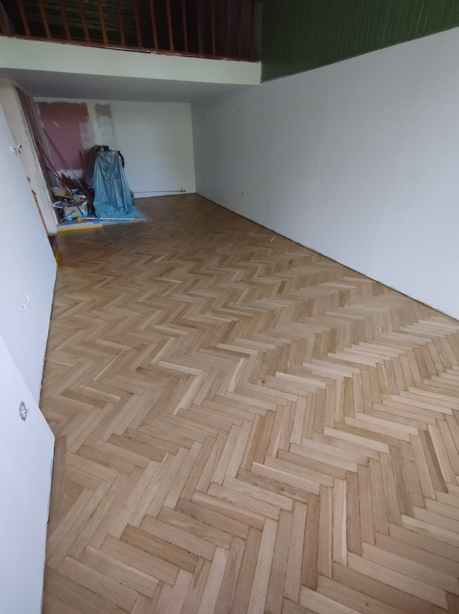 Ułożony parkiet w jodełkę w jasnym, przestronnym pokoju z przygotowanym miejscem do malowania ścian w tle.