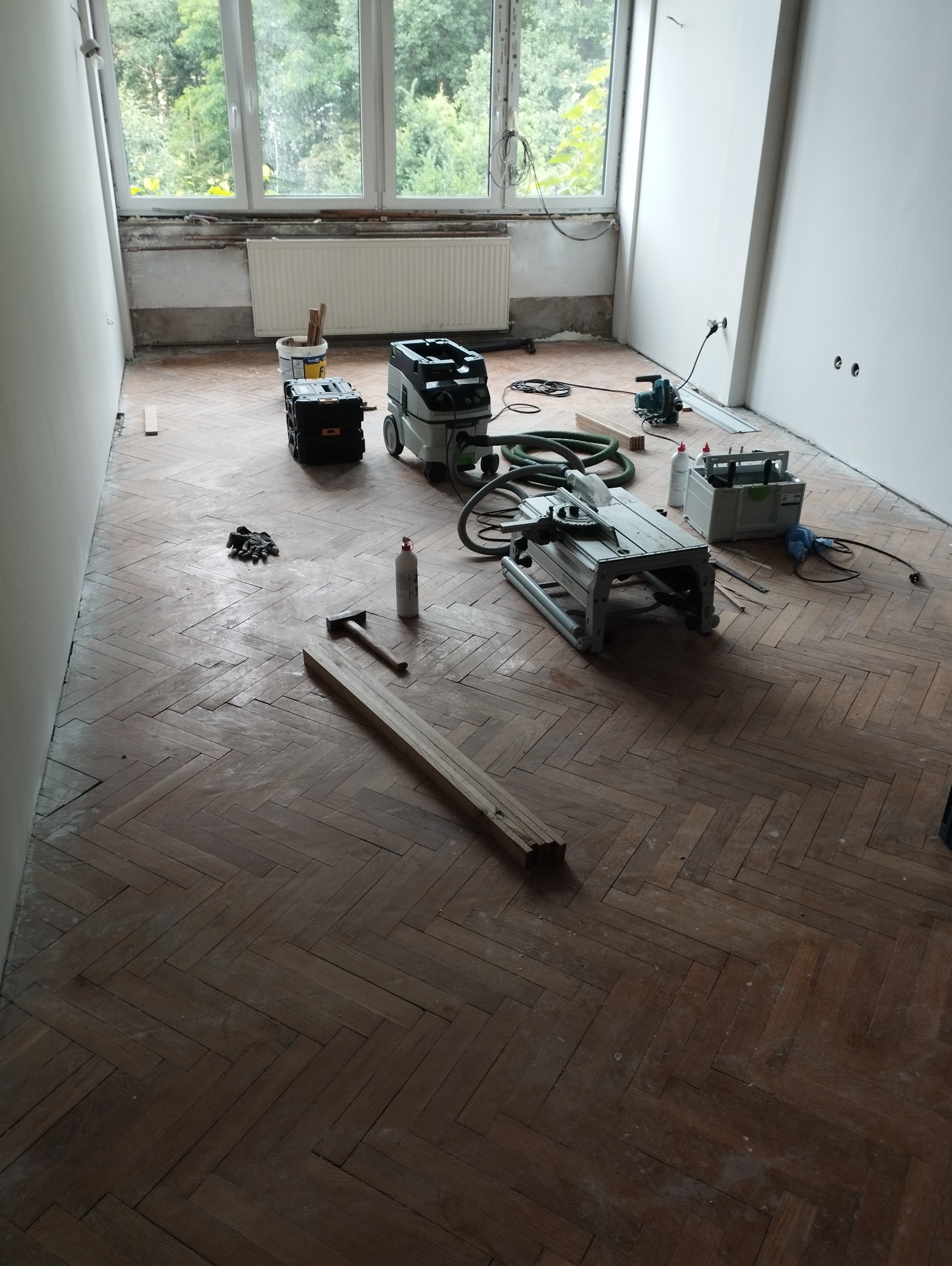 Pokój w trakcie renowacji podłogi z parkietu ułożonego w jodełkę, widoczne narzędzia: piła stołowa, odkurzacz przemysłowy, młotek, klej oraz deski. Na podłodze leżą robocze rękawice.