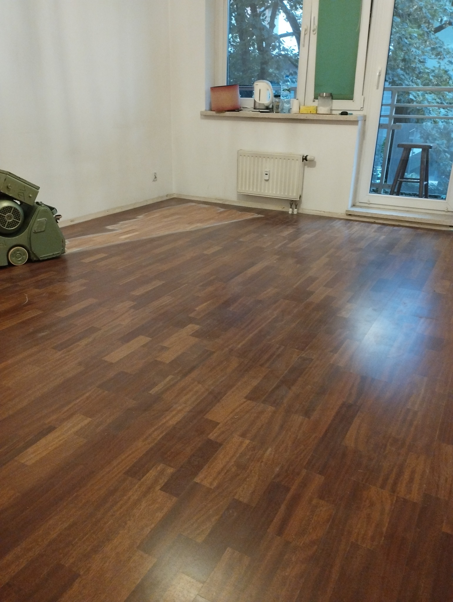 Częściowo odnowiona drewniana podłoga z parkietu w pokoju, z widoczną maszyną do szlifowania parkietu w lewym rogu i fragmentem podłogi przed cyklinowaniem przy ścianie.