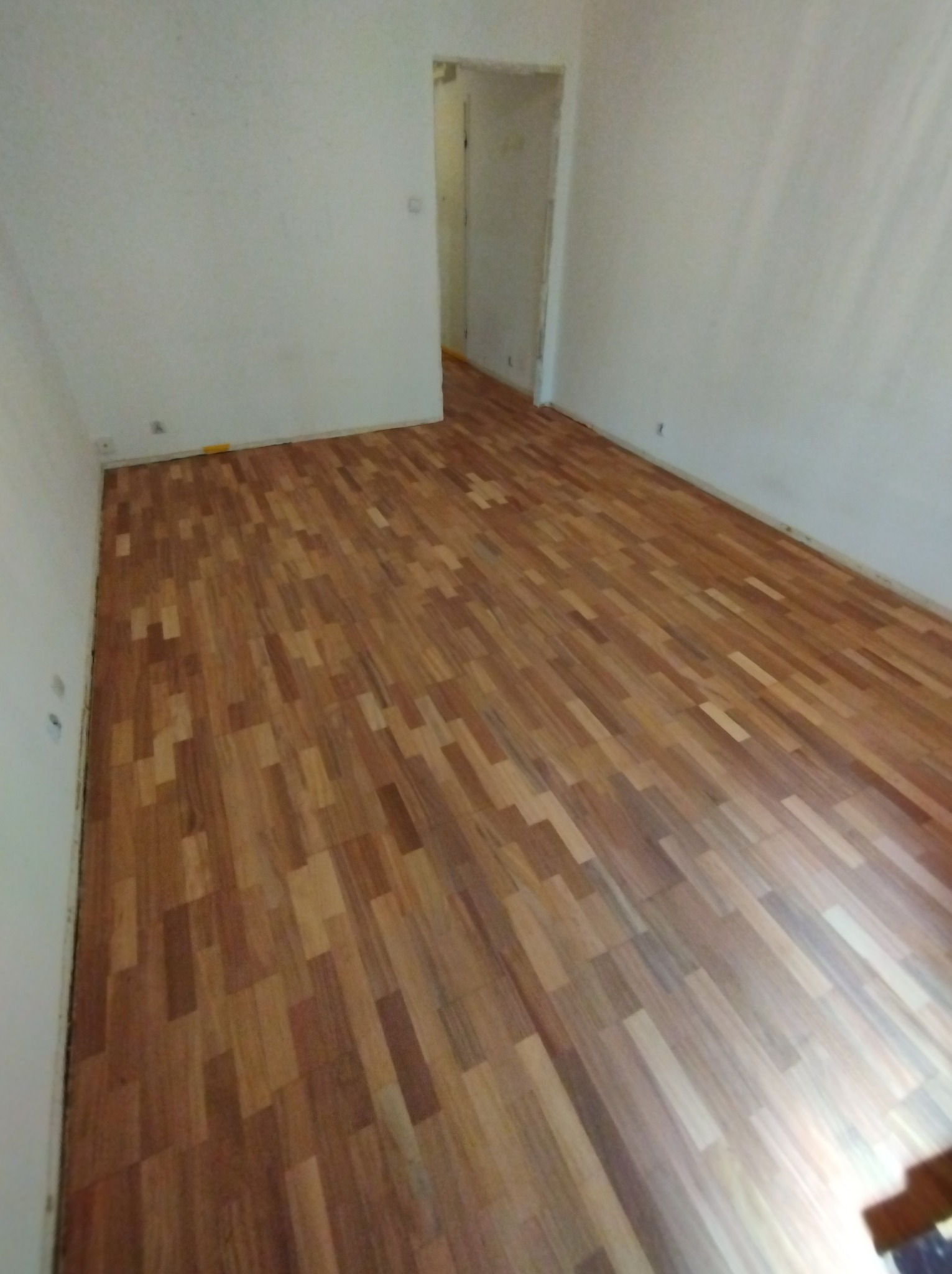 Wnętrze pokoju z nowo ułożonym parkietem w odcieniach brązu, widoczne białe ściany i przejście do innego pomieszczenia.