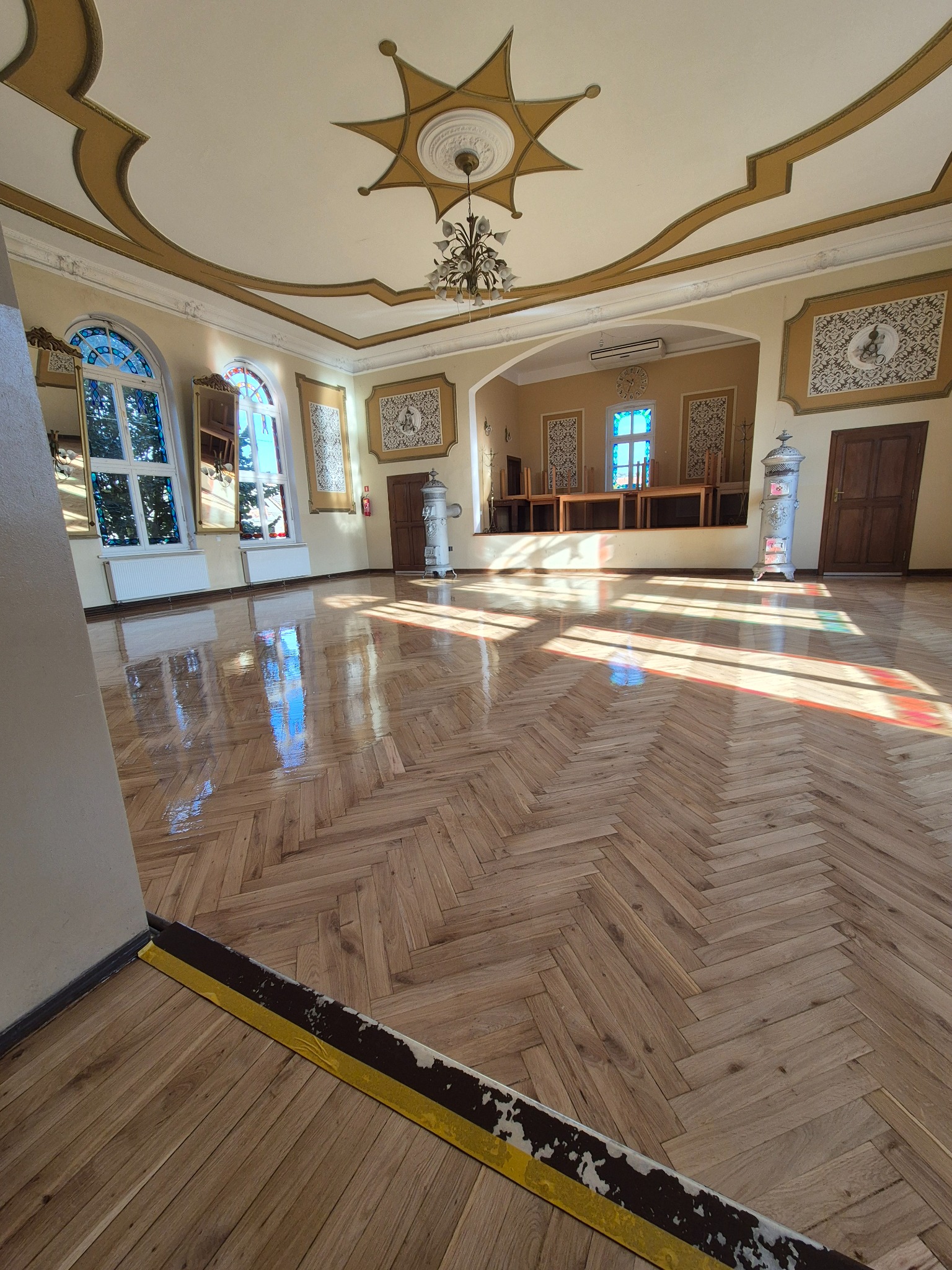 Sala z parkietem ułożonym w jodełkę, po cyklinowaniu i lakierowaniu, z widocznymi refleksami światła słonecznego, dekoracyjnym sufitem i pomalowanym progiem.