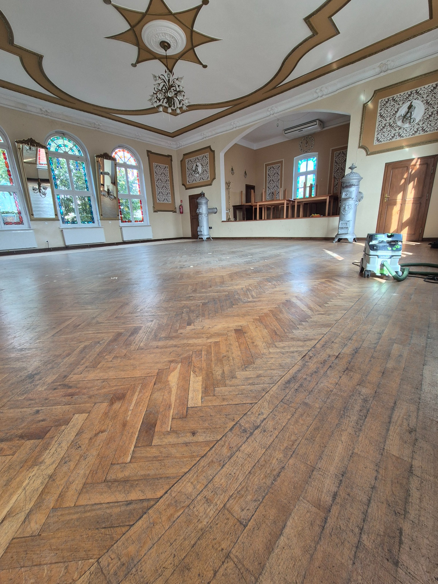 Sala balowa z parkietem ułożonym w jodełkę przed cyklinowaniem, widoczny odkurzacz przemysłowy i ozdobne piece kaflowe.