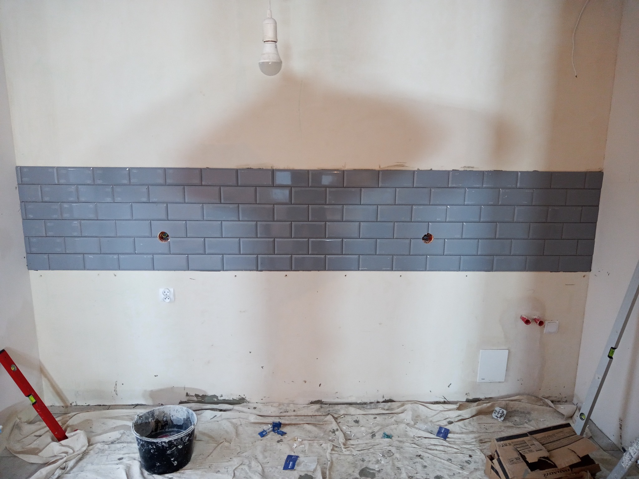 Ściana z częściowo ułożonymi szarymi, prostokątnymi płytkami ceramicznymi typu metro, z widocznymi otworami na gniazdka, wiadro i poziomica na podłodze zabezpieczonej folią malarską.