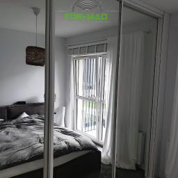 FOR-MAD Adrian Markiewicz - Szafa przesuwna z lustrzanymi drzwiami w sypialni, z odbiciem łóżka, okna balkonowego i lampy, na lustrze widoczne logo firmy FOR-MAD.