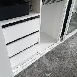 FOR-MAD Adrian Markiewicz - Wnętrze białej szafy przesuwnej z trzema szufladami, półką i wysuwanym wieszakiem na spodnie, widok z dołu na szarą wykładzinę.