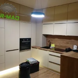 Nowoczesna kuchnia z beżowymi frontami szafek, drewnianym blatem i oświetleniem LED, z widocznym logo firmy na górnych szafkach.