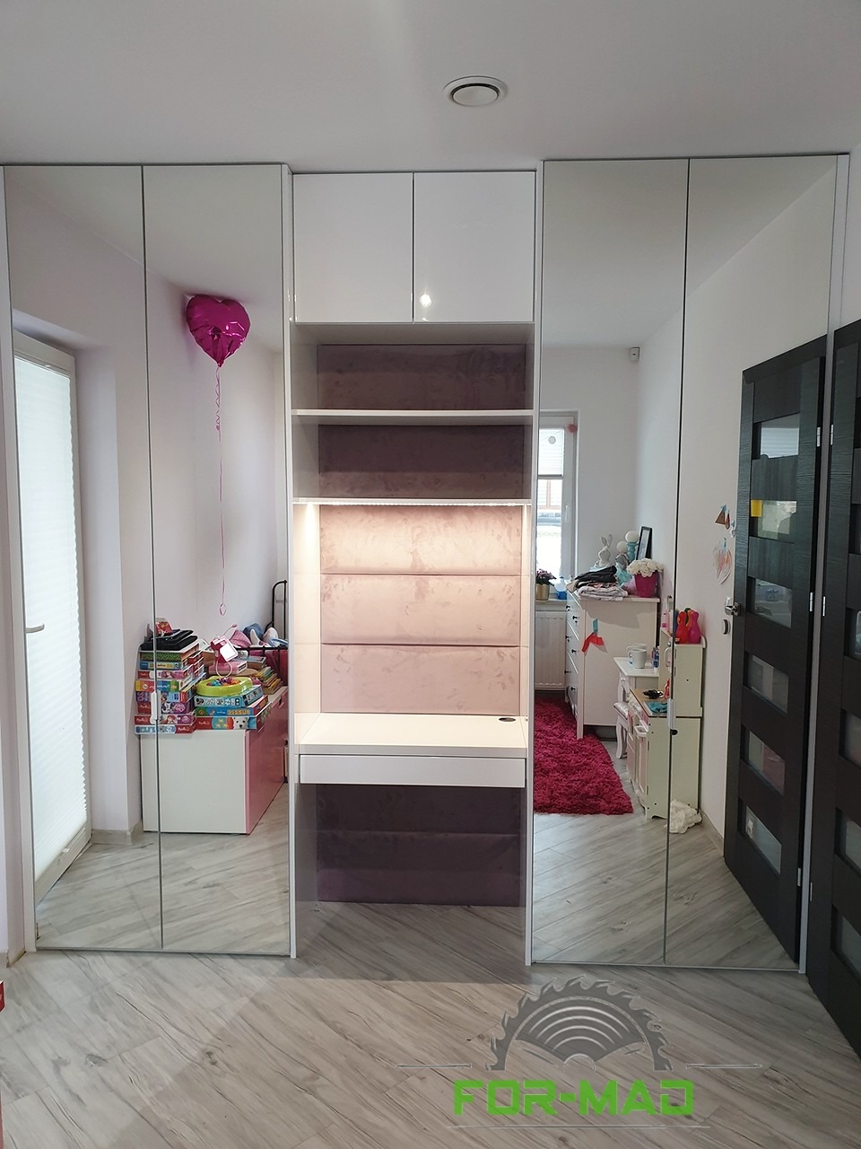 Pokój dziecięcy z szafą wnękową z lustrzanymi drzwiami i wbudowanym biurkiem z tapicerowanym panelem ściennym w odcieniach różu i szarości.