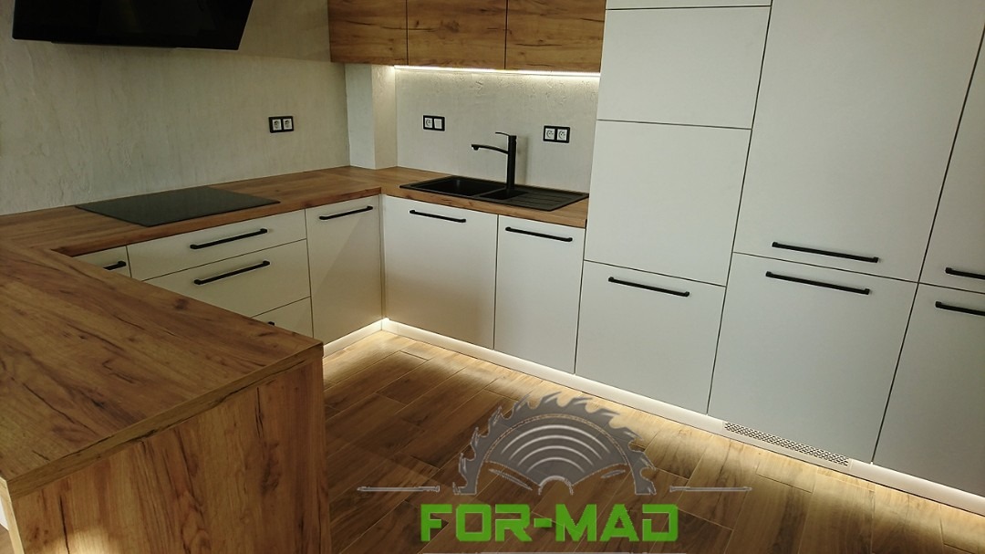 Nowoczesna kuchnia z białymi frontami i drewnianym blatem, oświetlenie LED pod szafkami, czarny zlew i bateria, widoczne gniazdka elektryczne na ścianie.