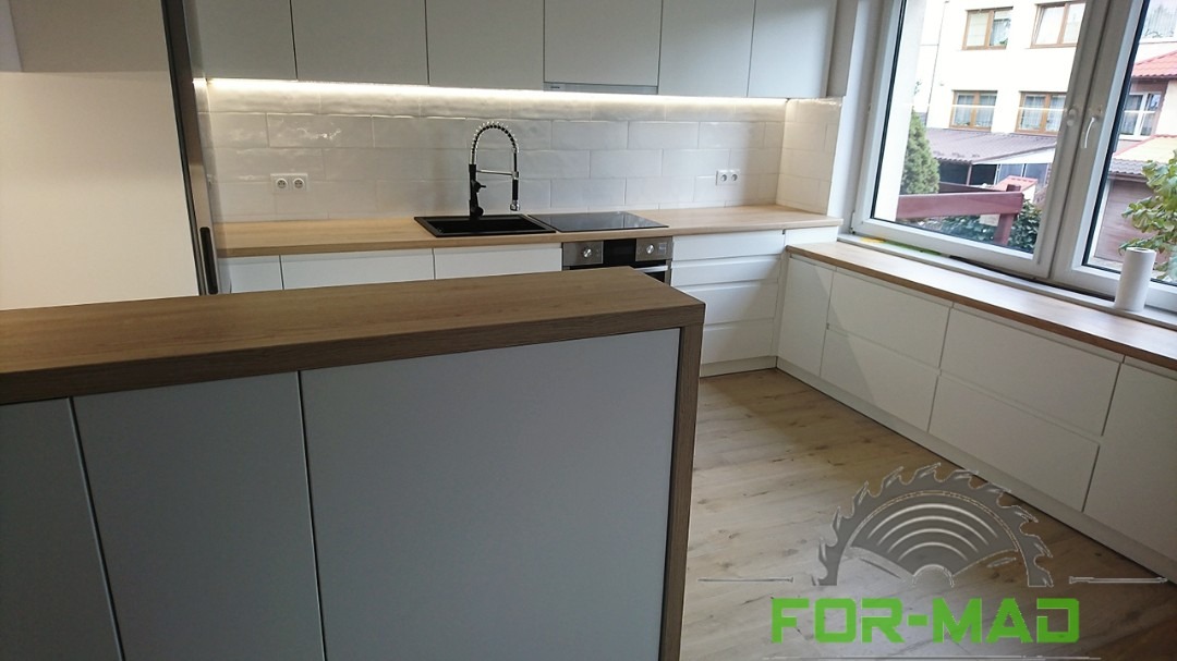 Biała kuchnia na wymiar z drewnianym blatem i wyspą kuchenną, widok z perspektywy podłogi, z widocznym logo firmy na dole.