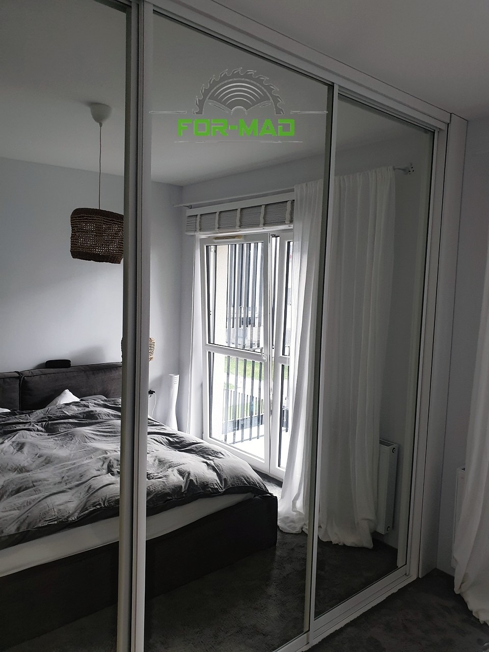 Szafa przesuwna z lustrzanymi drzwiami w sypialni, z odbiciem łóżka, okna balkonowego i lampy, na lustrze widoczne logo firmy FOR-MAD.