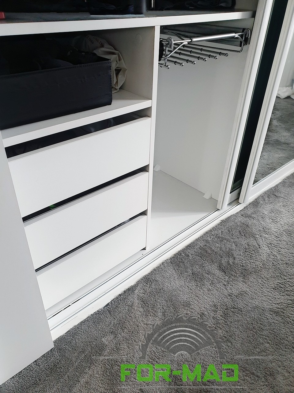 Wnętrze białej szafy przesuwnej z trzema szufladami, półką i wysuwanym wieszakiem na spodnie, widok z dołu na szarą wykładzinę.