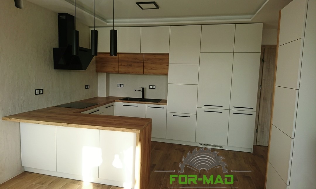 Nowoczesna kuchnia z białymi szafkami i drewnianym blatem, czarną armaturą i oświetleniem, widoczny fragment wyspy kuchennej.