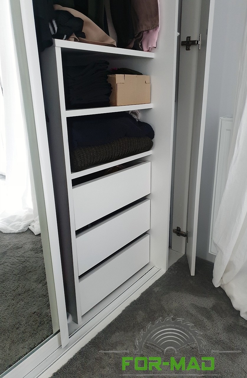 Wnętrze białej szafy z przesuwnymi drzwiami, widoczne półki z ubraniami i trzy szuflady, częściowo otwarte drzwi, na podłodze szary dywan, logo firmy w rogu.