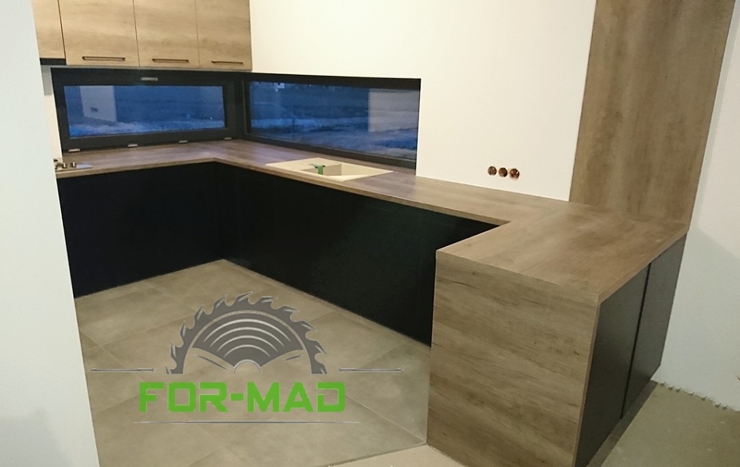 Nowoczesna kuchnia z blatem w kolorze drewna i czarnymi szafkami, szerokie okno nad blatem, widoczny logo firmy na podłodze, niedokończone wykończenie podłogi.