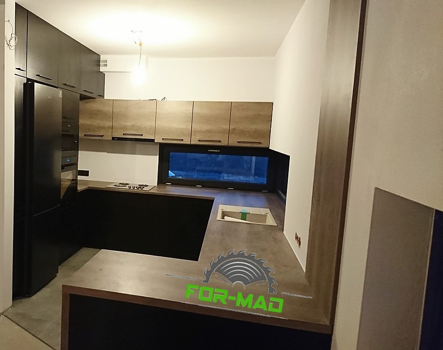 Nowoczesna kuchnia z ciemnymi szafkami dolnymi i jasnymi górnymi, z dużym oknem nad blatem, widoczne logo firmy na blacie.