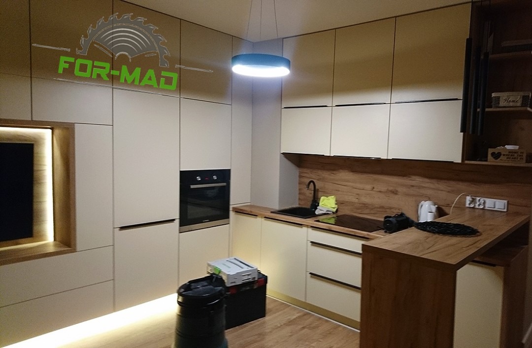 Nowoczesna kuchnia z beżowymi frontami szafek, drewnianym blatem i oświetleniem LED, z widocznym logo firmy na górnych szafkach.