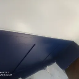 Pomieszczenie w trakcie wykańczania: granatowa, drewnopodobna boazeria ścienna z dekoracyjnymi wgłębieniami, biały sufit, zabezpieczona folią podłoga. Widoczny fragment ściany z naklejoną taśmą...