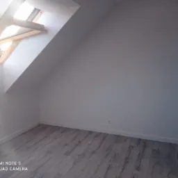 Poddasze po remoncie: świeżo pomalowane ściany w odcieniach bieli, drewniane okno dachowe z widokiem na drzewa bez liści, szare panele podłogowe imitujące drewno, listwy przypodłogowe, drzwi...