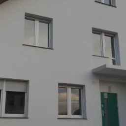 Dwupiętrowy budynek z oknami i zielonymi drzwiami po świeżym malowaniu elewacji na biało, widoczne odbicie nieba w szybach okiennych.