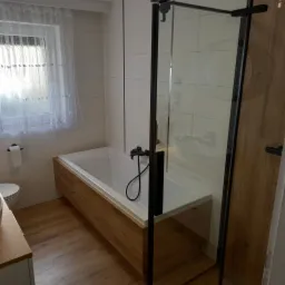 Nowoczesna łazienka z wanną obudowaną drewnem, czarną armaturą i szklanym prysznicem typu walk-in, wykończona w jasnych płytkach, z oknem z firanką.