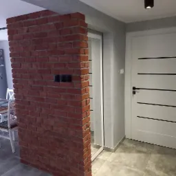 Ceglana ścianka działowa z widocznymi gniazdkami elektrycznymi, przylegająca do ściany z drzwiami w nowoczesnym wnętrzu.