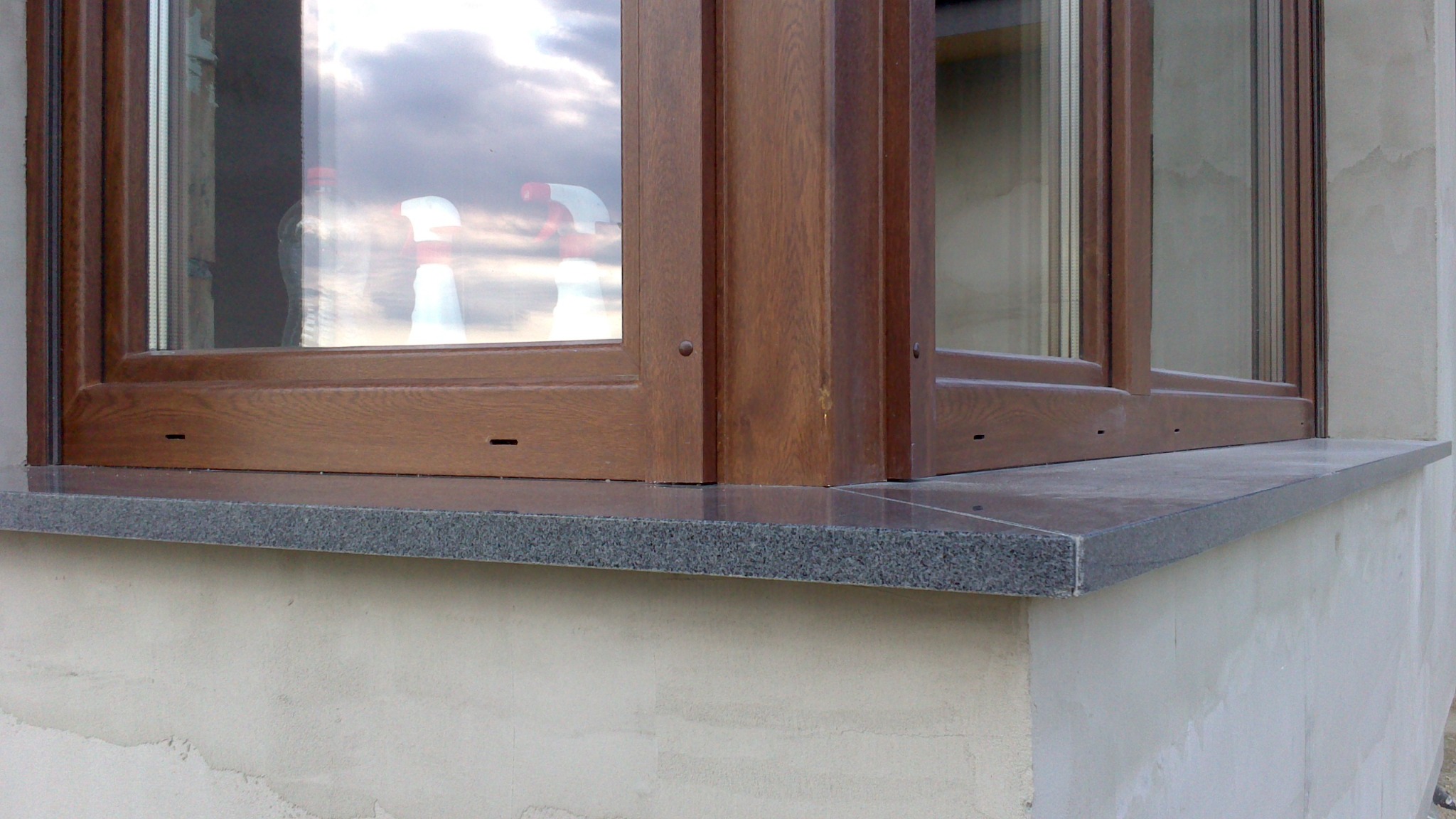 Zamontowany, ciemnoszary parapet z granitu pod brązowym oknem PCV, widoczny fragment surowej elewacji budynku.