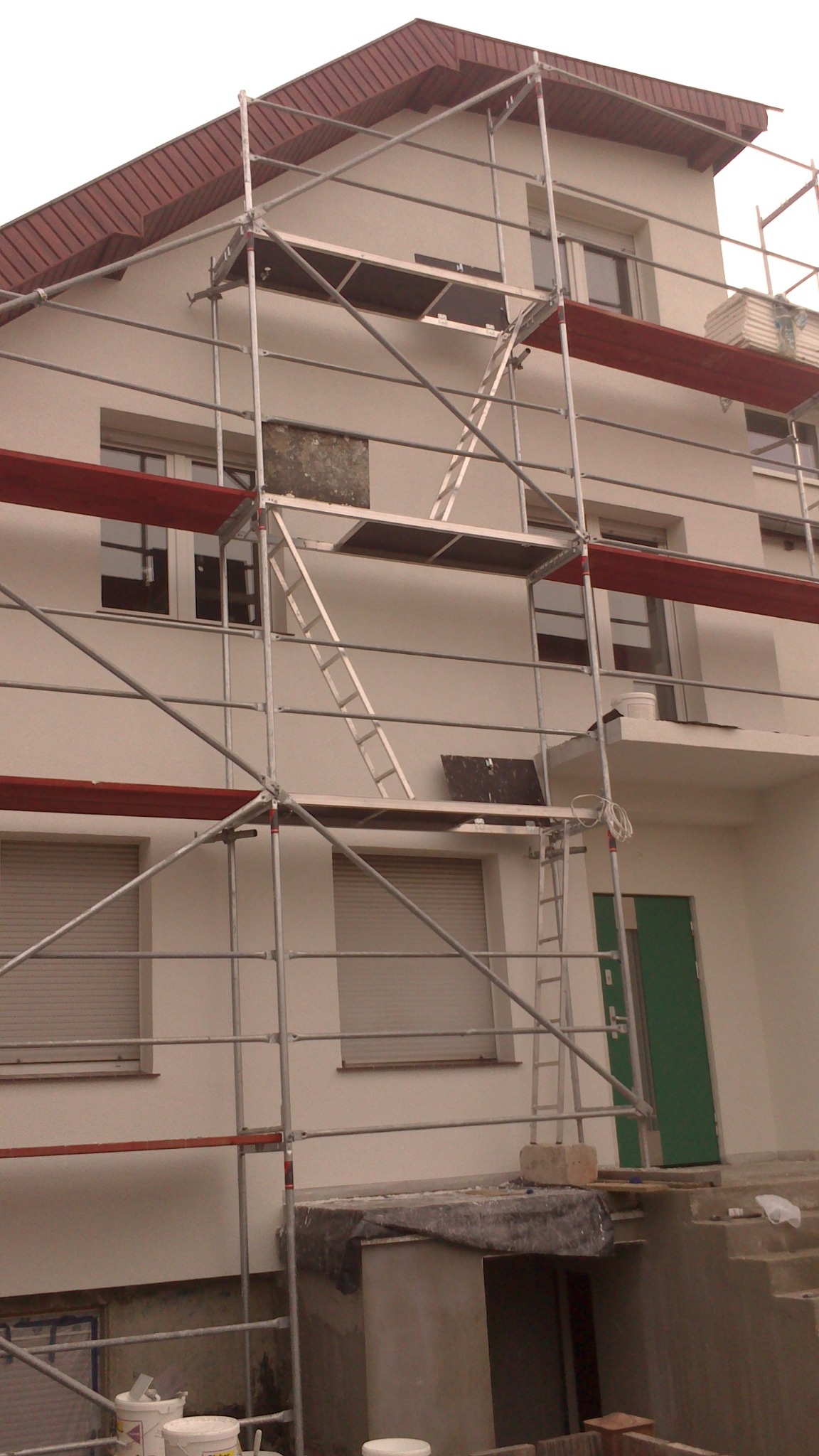 Dom w trakcie prac elewacyjnych, widoczne rusztowanie, odkryty fragment ściany i pojemniki z farbą u podstawy.