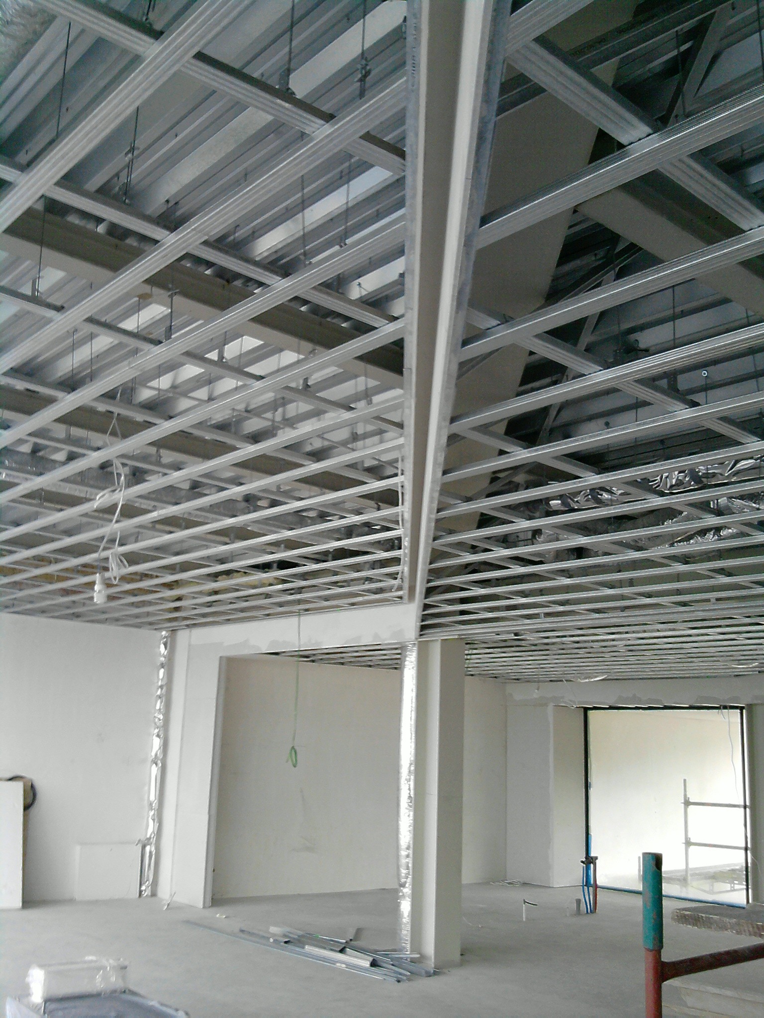 Montaż metalowej konstrukcji sufitu podwieszanego z płyt kartonowo-gipsowych, widoczne profile, zawiesia i częściowo zamontowana izolacja, w tle białe ściany i okno.