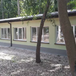 Jasnozielony budynek z białymi oknami w otoczeniu drzew, widok z zewnątrz, ziemia pokryta liśćmi i gałęziami.