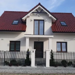 Nowoczesny dom jednorodzinny z ciemną stolarką okienną i czerwoną dachówką, widok z zewnątrz przez metalowe ogrodzenie.