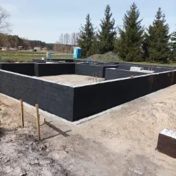 Wzniesione ściany fundamentowe z bloczków pomalowane na czarno, widoczne zbrojenia i szalunki, w tle zielony teren z drzewami i toaleta przenośna.