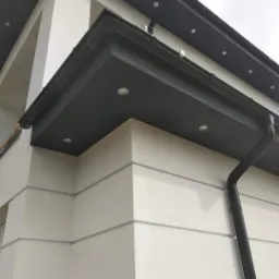 Fragment elewacji budynku z widocznymi elementami dekoracyjnymi, oświetleniem zewnętrznym i systemem rynnowym w kolorze grafitowym, z bliska.