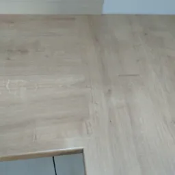 Fragment drewnianej podłogi z widocznym łączeniem paneli i fragmentem ościeżnicy drzwiowej w dolnym lewym rogu, ukazujący wnętrze z białymi płytkami.