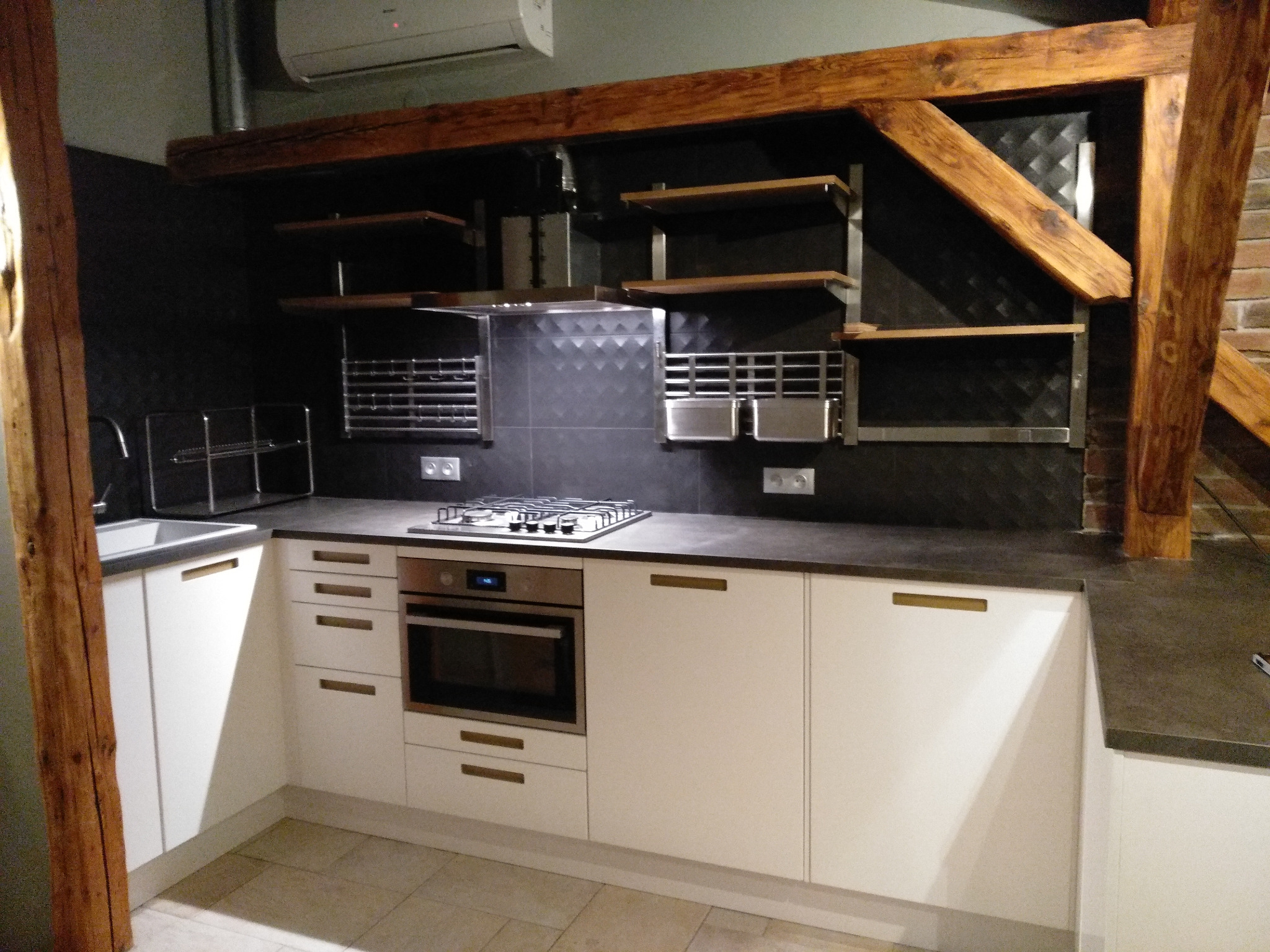 Nowoczesna kuchnia z białymi szafkami, ciemnym blatem i drewnianymi belkami stropowymi, oświetlona z góry, z widoczną płytą gazową i piekarnikiem w zabudowie.