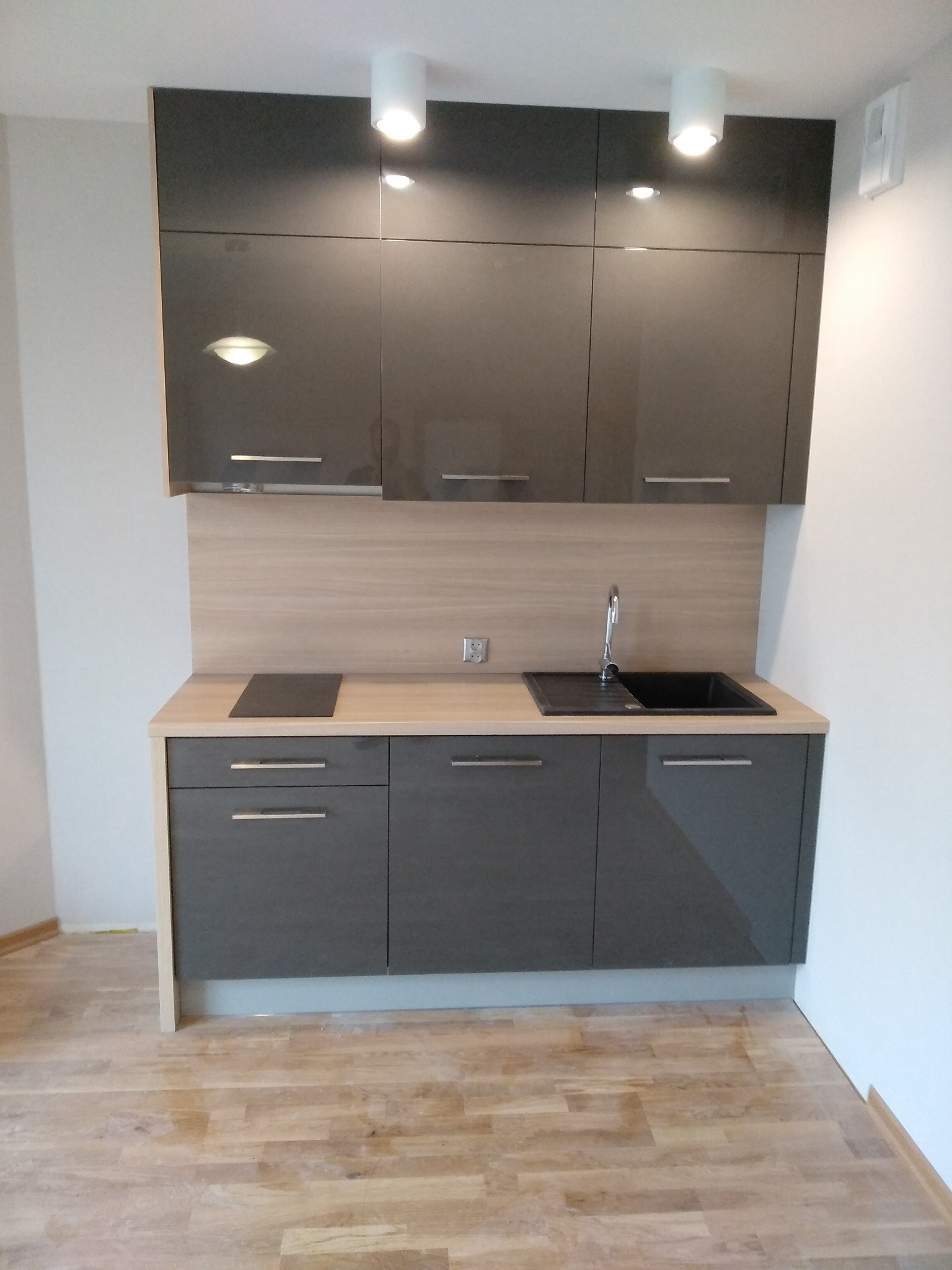 Szara, lakierowana zabudowa kuchenna z drewnianym blatem i płytkami imitującymi drewno, oświetlona trzema lampami sufitowymi.