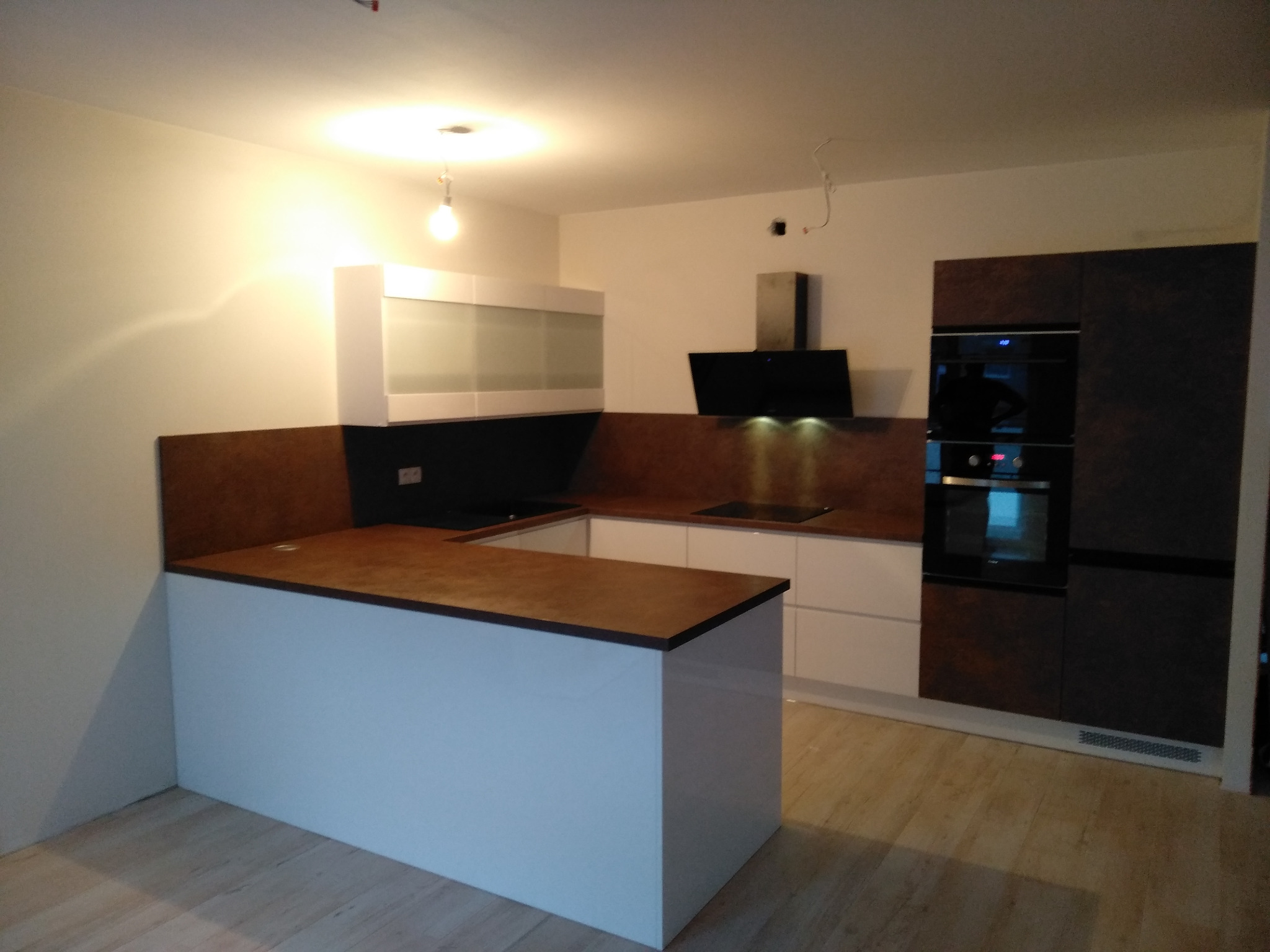 Nowoczesna kuchnia z białymi szafkami, blatem w kolorze ciemnego drewna i wyspą kuchenną, oświetlona wiszącą lampą, z widocznym okapem i zabudowanym piekarnikiem.