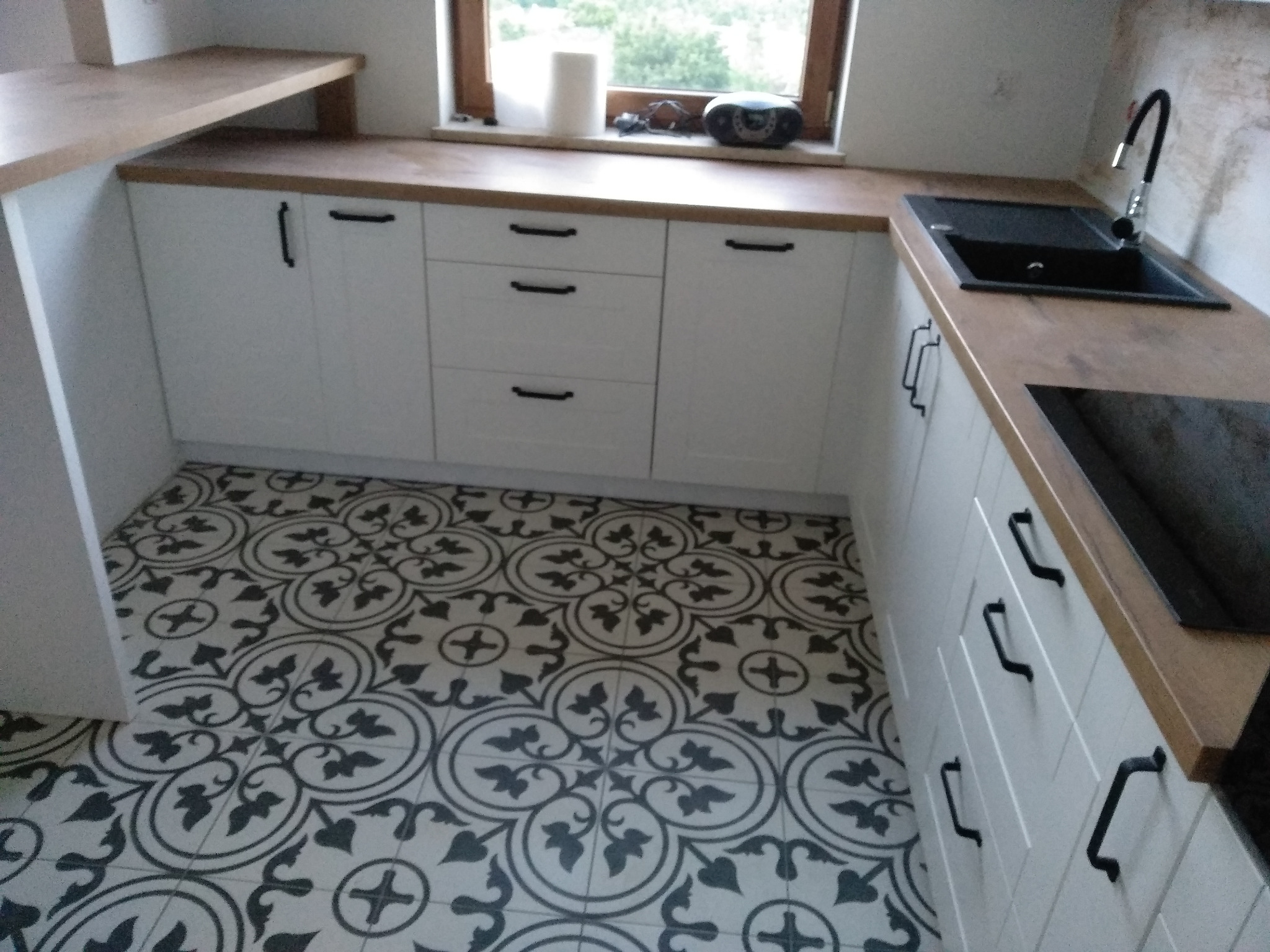 Nowoczesna zabudowa kuchenna z białymi szafkami i blatem imitującym drewno, czarny zlew i bateria, wzorzysta podłoga w stylu retro.