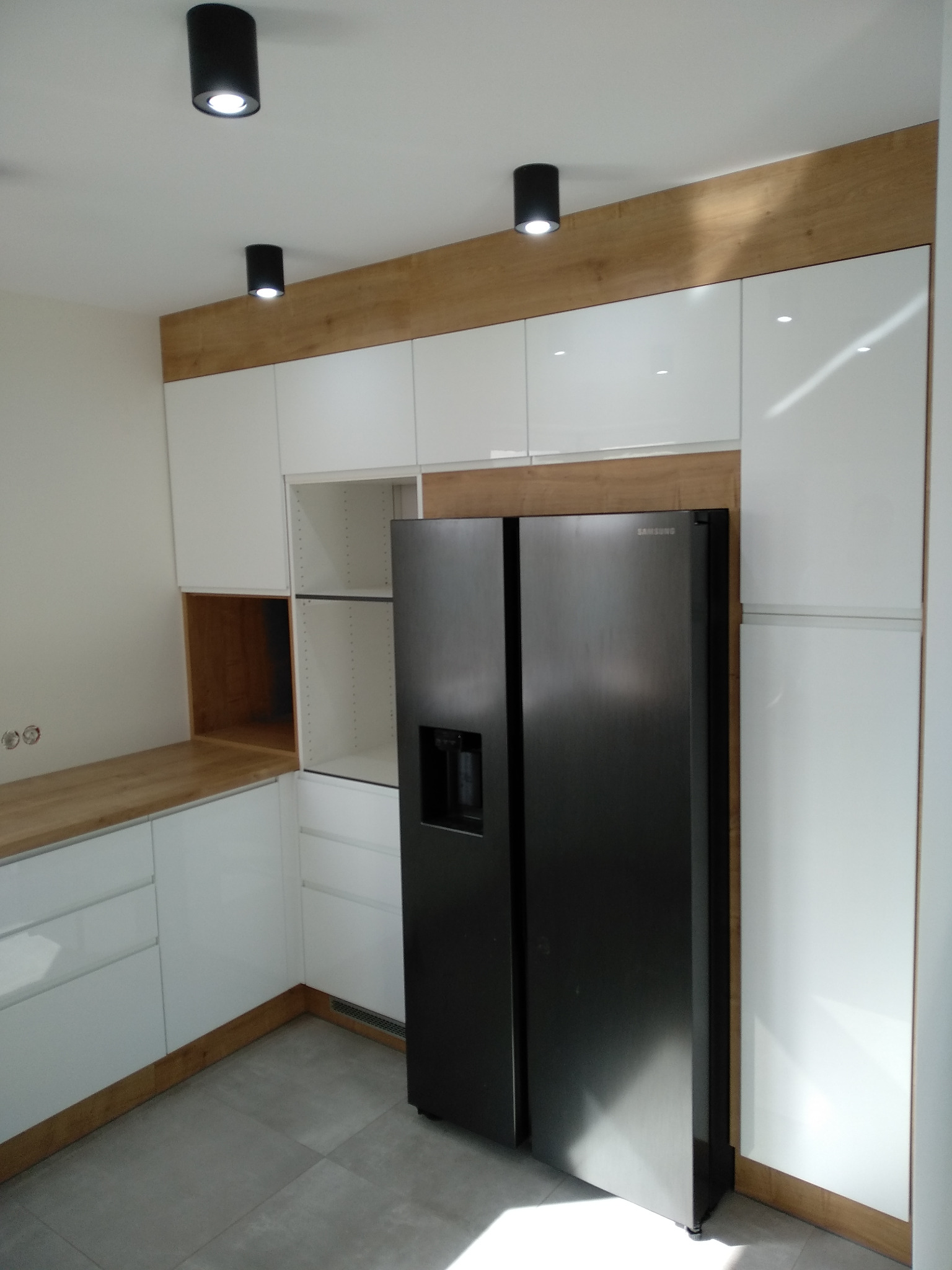 Nowoczesna kuchnia z białymi, lakierowanymi szafkami, drewnianymi akcentami i czarną, dwudrzwiową lodówką Samsung z dystrybutorem wody.