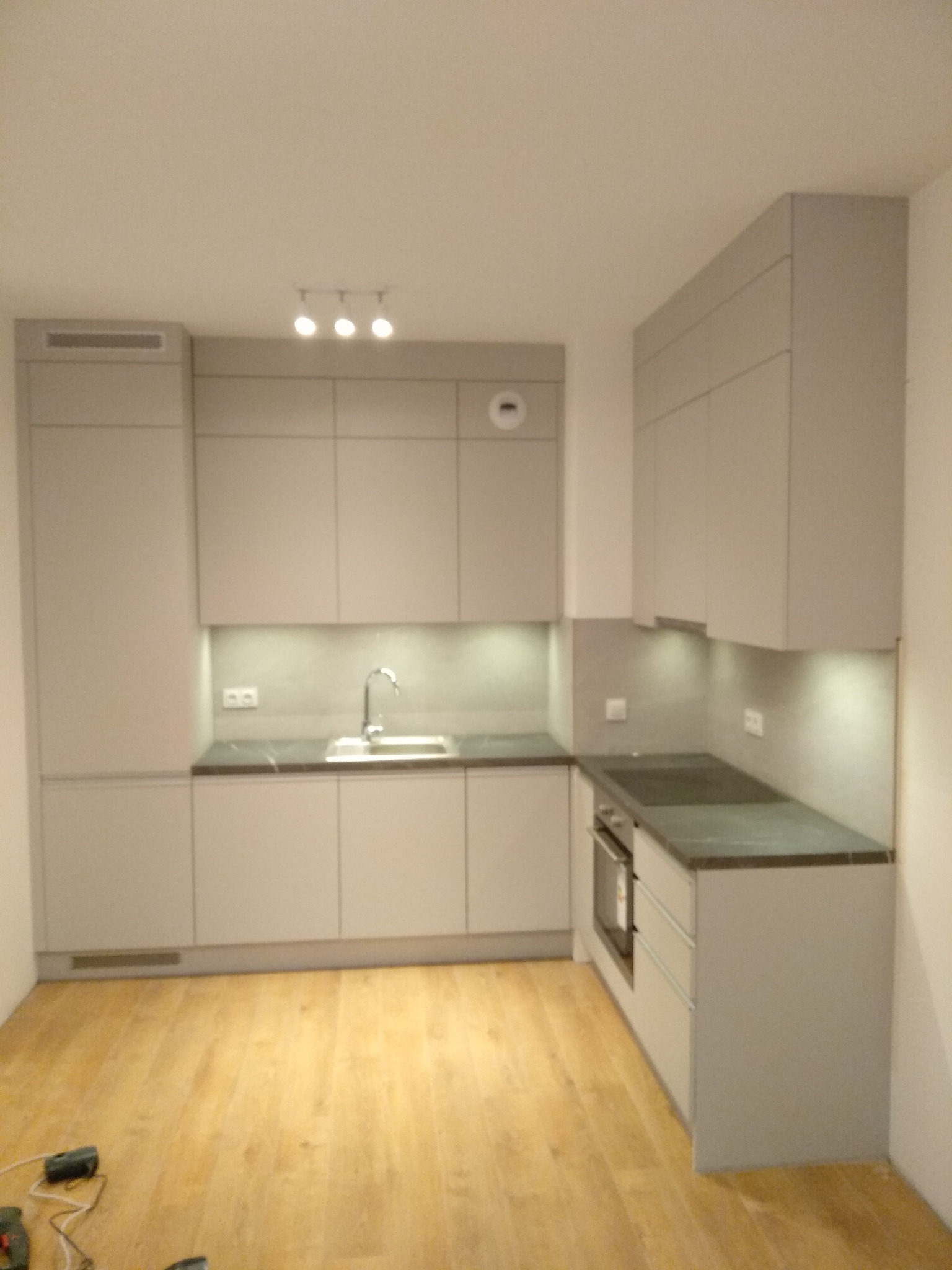 Nowoczesna kuchnia w odcieniach szarości z drewnianą podłogą, oświetleniem podszafkowym i trzema lampami sufitowymi.