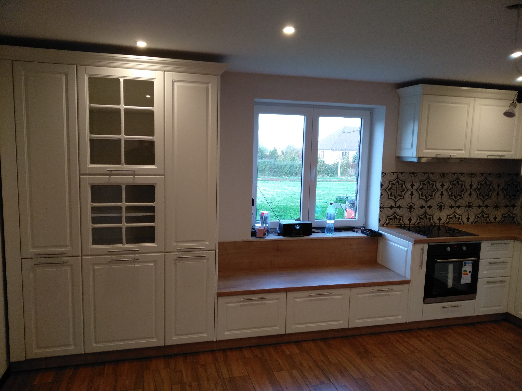 Biała zabudowa kuchenna z drewnianym blatem i przeszklonymi szafkami w Krakowie, z widokiem na ogród przez okno.