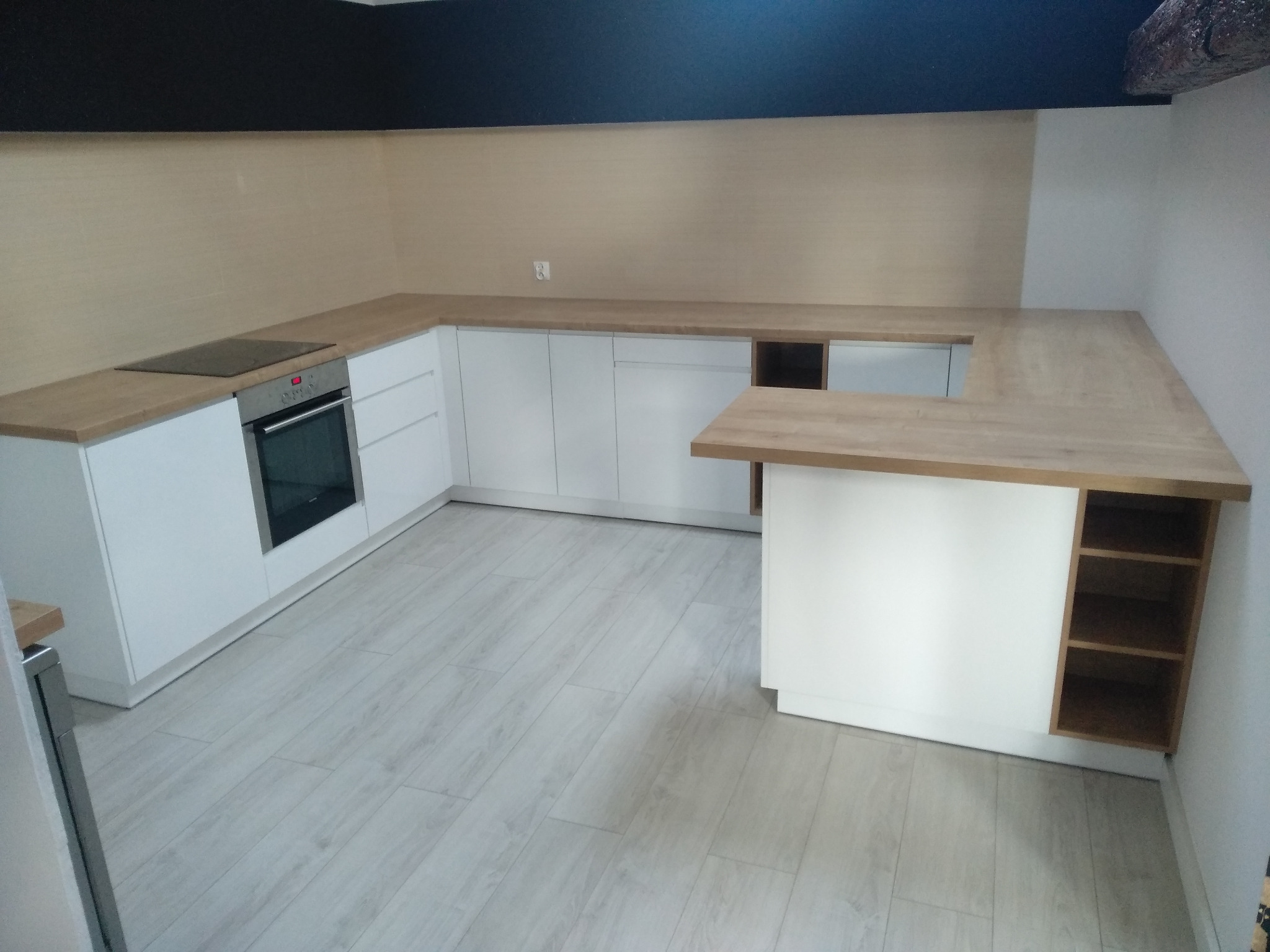 Nowoczesna kuchnia z białymi szafkami i blatem z jasnego drewna, widok na wyspę kuchenną z półkami, płyta indukcyjna i piekarnik w zabudowie, szare panele podłogowe.